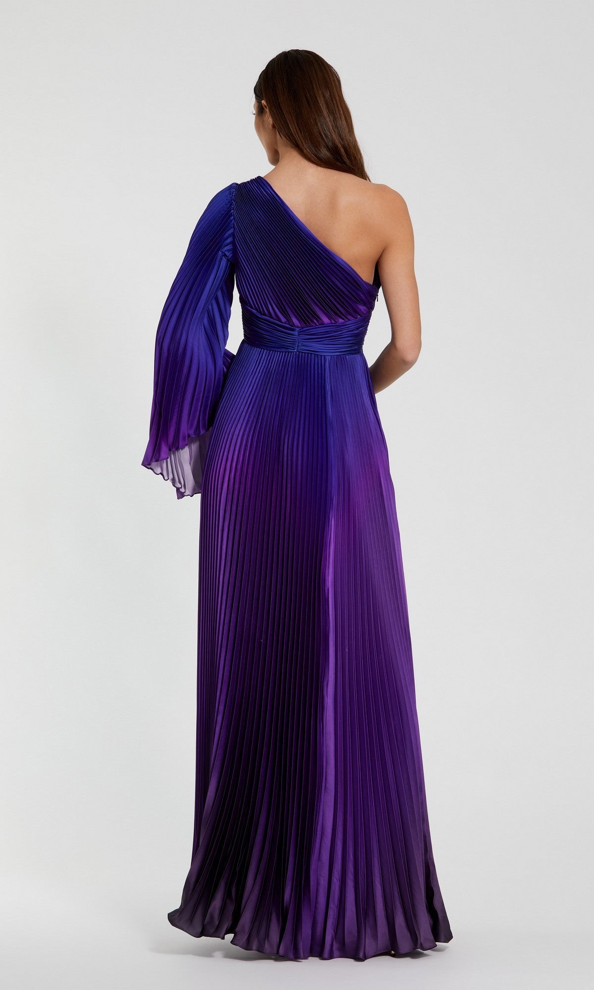 A woman stands with her back to the camera in a Mac Duggal 49903 One-Shoulder Ombre Formal Dress, featuring floor-length pleats in purple and blue against a plain background.